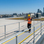 Un technicien portant un casque et un gilet de haute visibilité inspecte une passerelle sécurisée sur un toit urbain.